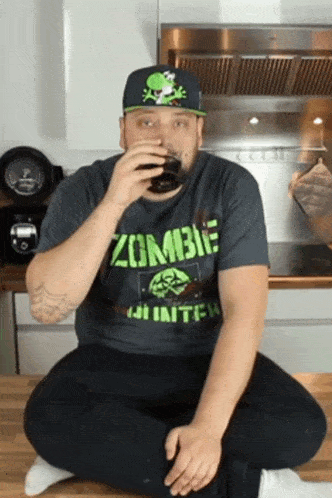 a man wearing a zombie shirt sits on the floor drinking a glass of soda