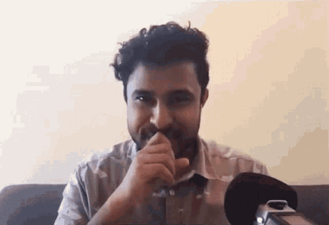 a man with a beard is sitting on a couch and covering his mouth with his hands .