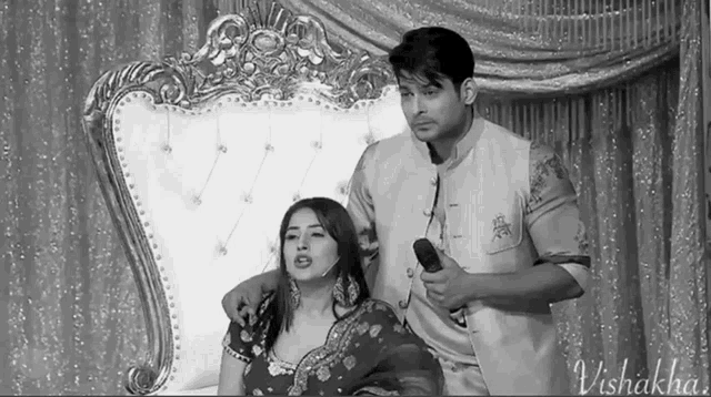 a black and white photo of a man holding a microphone next to a woman sitting on a couch .