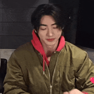 a man wearing a green jacket and a red hoodie is sitting at a table with his eyes closed .