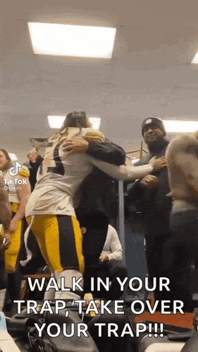 a football player is hugging another player in a locker room while a group of people watch .