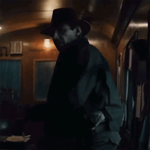 a man wearing a hat and a brown coat stands in a dark room