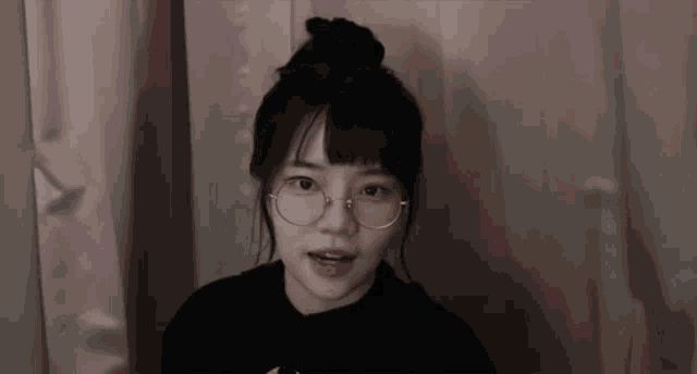 a young woman wearing glasses and a black sweater is standing in front of a curtain .