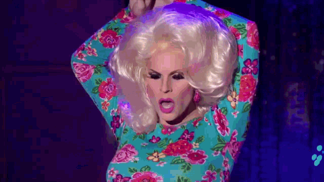 a drag queen is wearing a floral dress and wig and dancing on a stage .