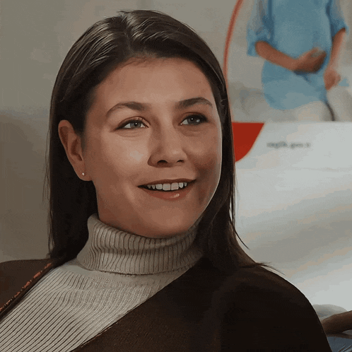 a woman wearing a turtleneck sweater smiles in front of a sign that says ' sağlık ' on it