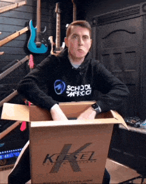 a man wearing a hoodie that says school of music sits in a cardboard box