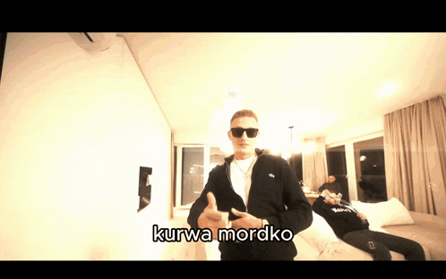 a man wearing sunglasses stands in a room with kurwa mordko written on the bottom right