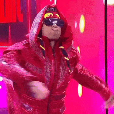 a man in a red hooded jacket and sunglasses stands in front of a pink background