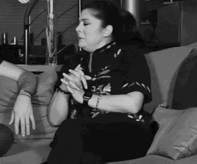 a black and white photo of a woman sitting on a couch with her hands folded .
