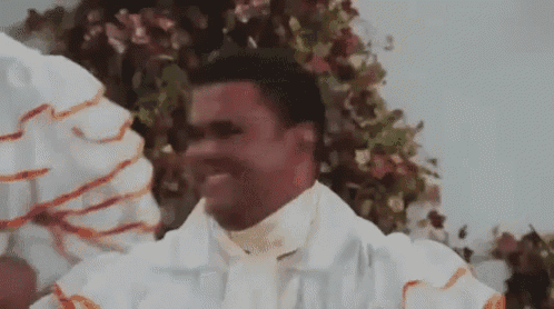 a man in a white robe is standing in front of a flower arrangement and smiling .