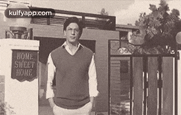 a man is standing in front of a house with his hands in his pockets and a sign that says `` home sweet home '' .