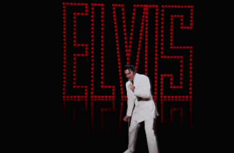 a man in a white suit singing into a microphone in front of a elvis sign