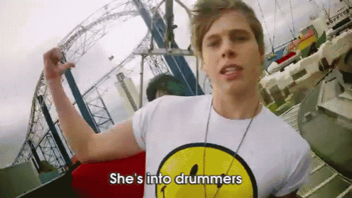 a man wearing a smiley face shirt says she 's into drummers in front of a roller coaster
