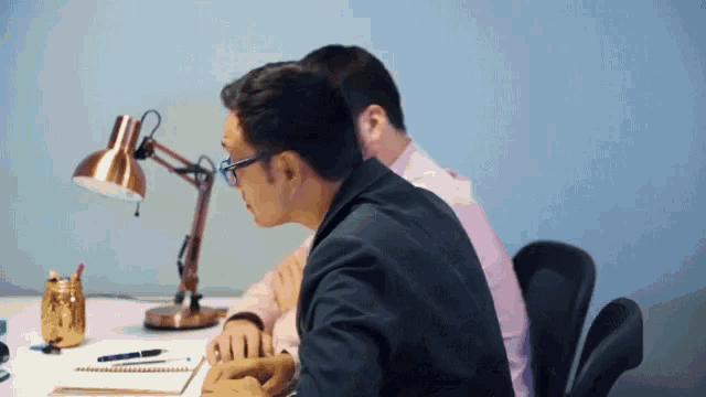 two men are sitting at a desk with a lamp on it