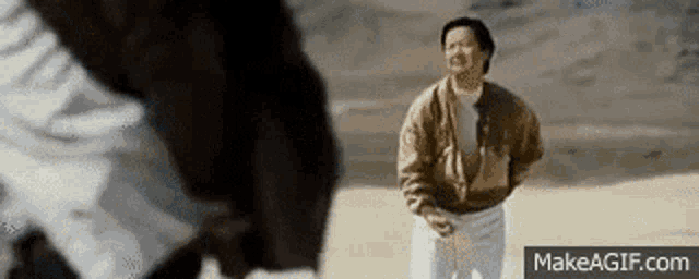 a man in a brown jacket is standing in the sand looking at another man in a white shirt .