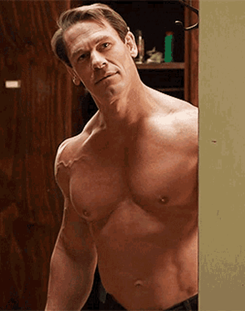 a shirtless man leans against a wall looking at the camera