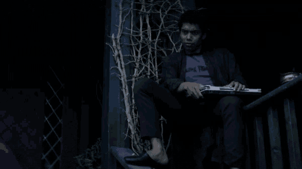 a man in a purple shirt is sitting on a porch holding a laptop and the word spooky is next to him