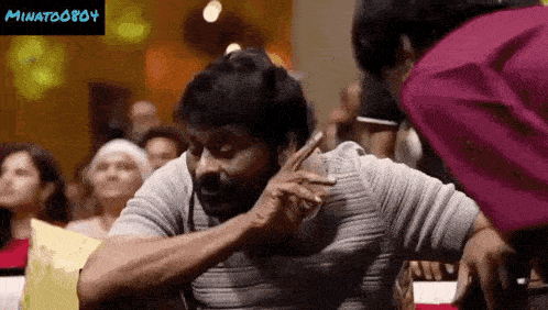 a man with a beard is sitting in front of a crowd and a woman is standing next to him .