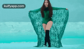 a woman in a green bodysuit and black boots is standing in front of a blue ocean .