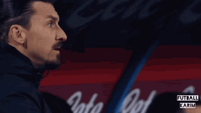 a man in a black jacket is clapping his hands in front of a sign that says ' futball karim '