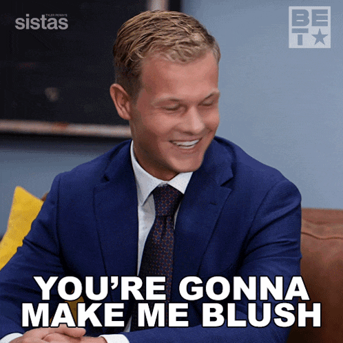 a man in a suit and tie is sitting on a couch and says you 're gonna make me blush