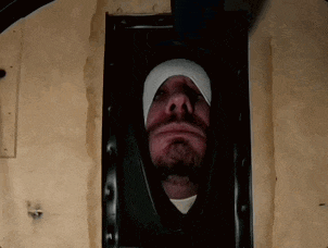 a man with a beard and a bandage on his head is looking out of a hole in a wall .