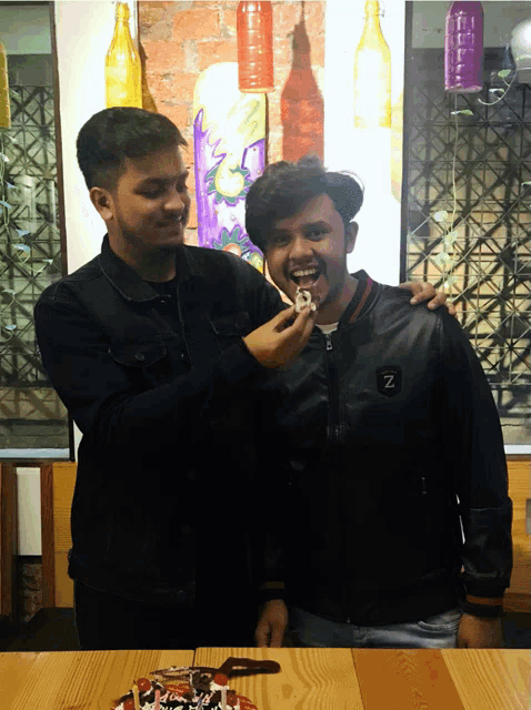 a man wearing a black jacket with the letter z on it feeds another man a piece of cake