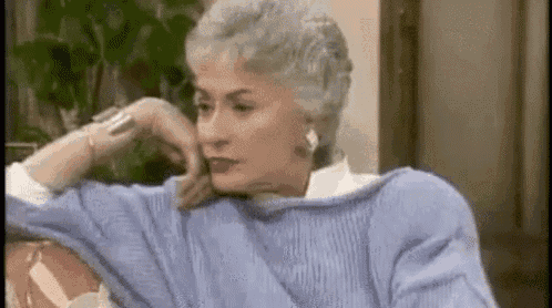 an older woman in a blue sweater is sitting on a couch with her hand on her chin .
