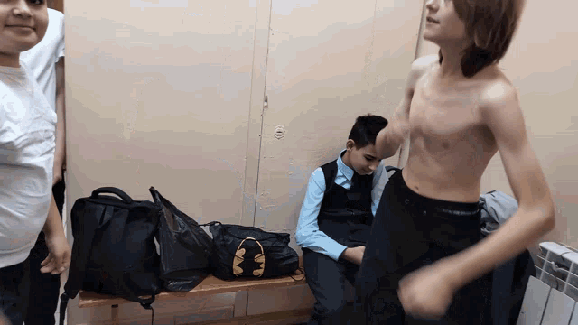 a boy without a shirt sits on a bench next to a black bag that says ' headphones ' on it