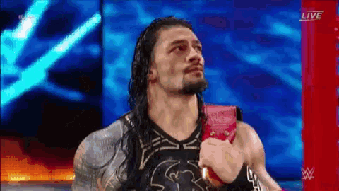 roman reigns is wearing a red belt on his shoulder while standing on a stage .