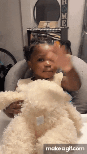 a little girl is holding a teddy bear and waving at the camera ..