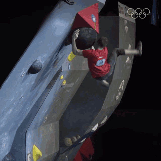 a man in a red shirt is climbing up a wall with the olympic rings in the background
