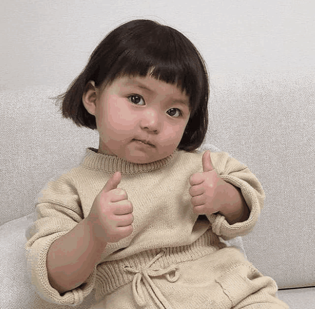 a little girl is sitting on a couch and giving a thumbs up .