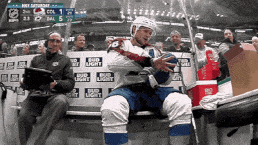 a hockey player sits on the bench during a game with bud light advertisements behind him