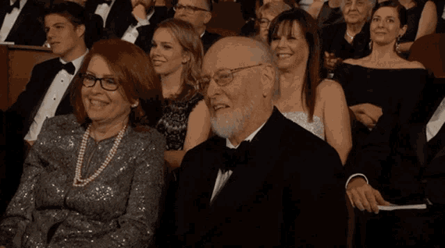 a man in a tuxedo and glasses sits in a crowd of people