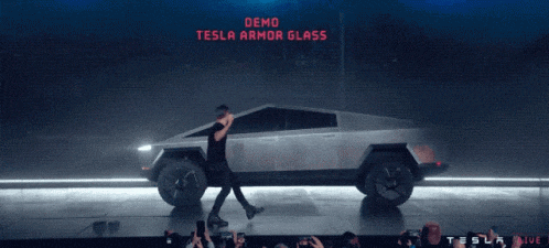 a man stands in front of a tesla truck with a demo tesla armor glass sign above him