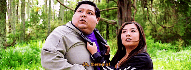 a man and a woman are standing next to each other in a forest and the woman is holding the man 's jacket .