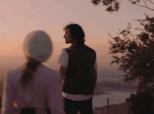 a man and a woman standing on top of a hill looking at the sunset