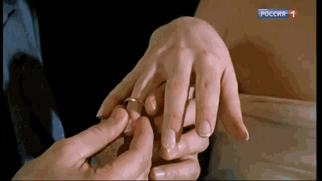 a man putting a wedding ring on a woman 's finger with a russian tv channel in the corner