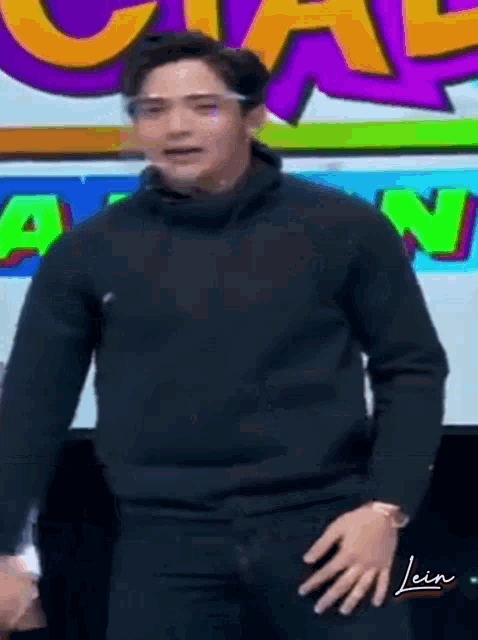 a man wearing a black sweater and glasses is standing in front of a screen with the letter n on it .