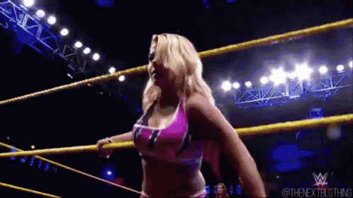 a woman in a pink top is standing in a wrestling ring with a yellow rope .