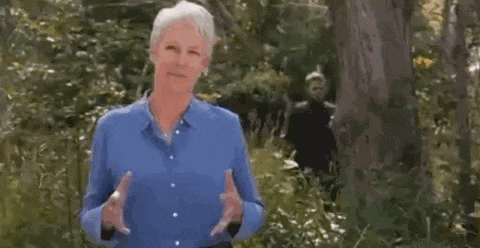 a woman in a blue shirt is standing next to a tree in the woods holding a cell phone .
