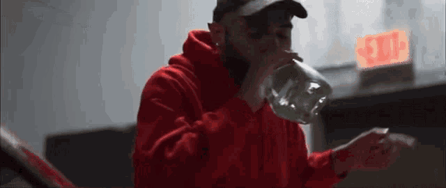 a man in a red hoodie is drinking from a bottle while standing in front of an exit sign .