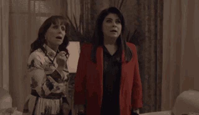 two women are standing next to each other in a living room and one of them is wearing a red jacket .