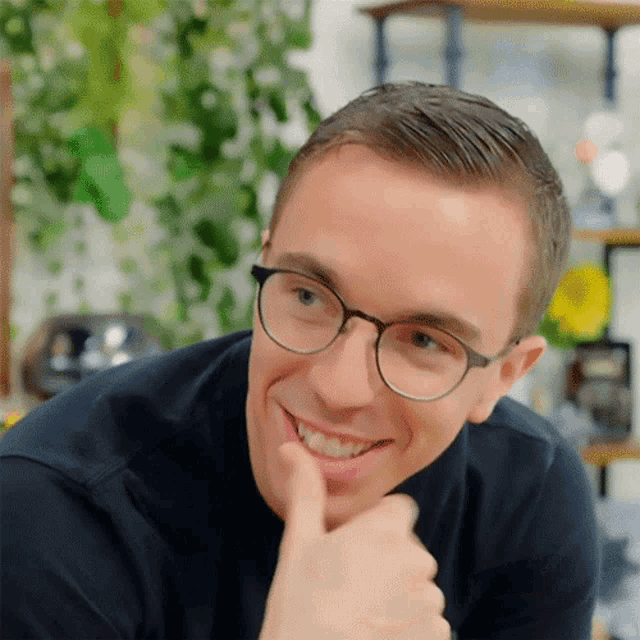 a man wearing glasses and a black shirt smiles with his finger on his chin