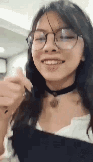 a young woman wearing glasses and a choker is smiling and making a heart shape with her hands .