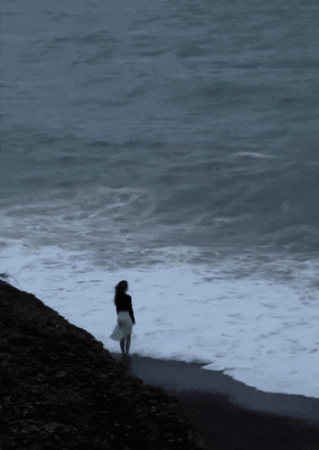 a woman in a white skirt is standing on the shore of a body of water