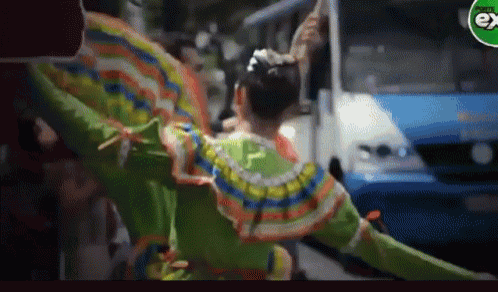a woman in a colorful dress is dancing in front of a bus with the word ex on the corner