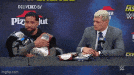 two men are sitting at a table with a pizza hut sign behind them
