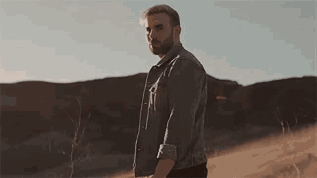a man in a denim jacket is standing in a field with a mountain in the background .